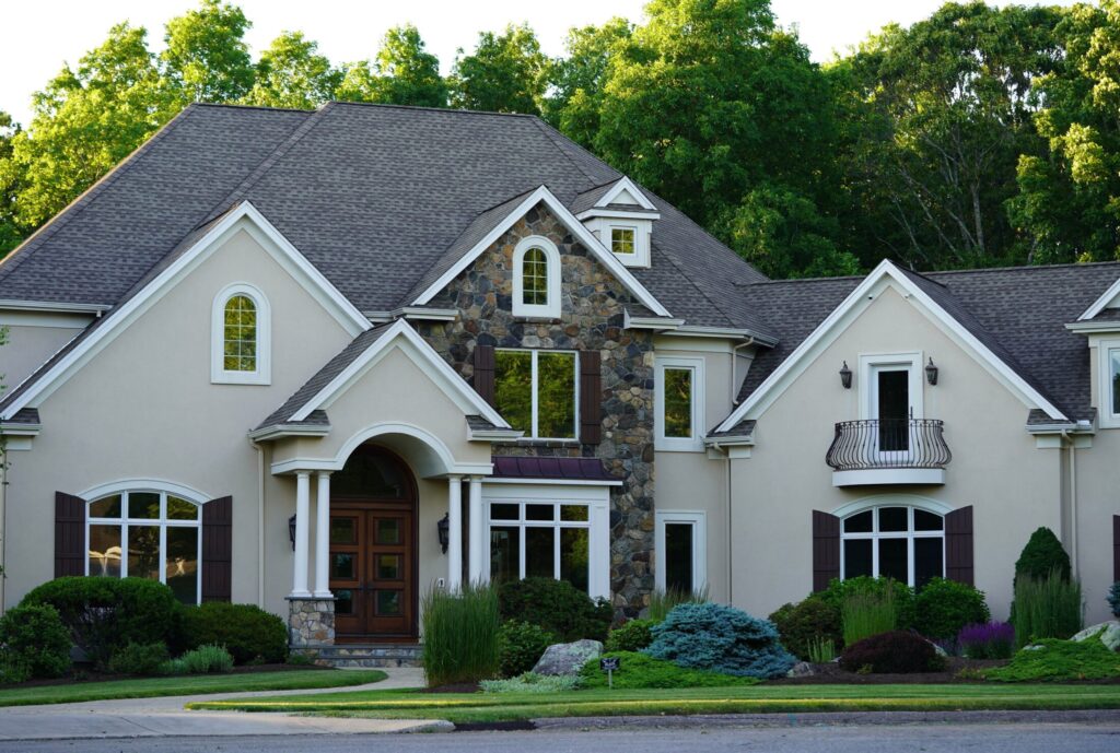 a large beige luxury home features a distinctive stone masonry accent dark window shutters a wrought-iron balcony and a meticulously landscaped front yard backed by dense green trees